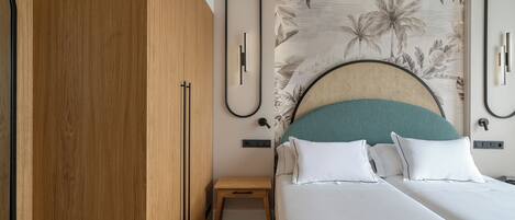 Un dormitorio moderno con un armario de madera, una cama con ropa de cama blanca y almohadas turquesas, una mesita de noche de madera pequeña, y una pared con un estampado de hojas tropicales.