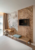 A modern hotel room with a large bed, a desk with a chair, a TV mounted on a bamboo-patterned wall, and a large window with curtains.