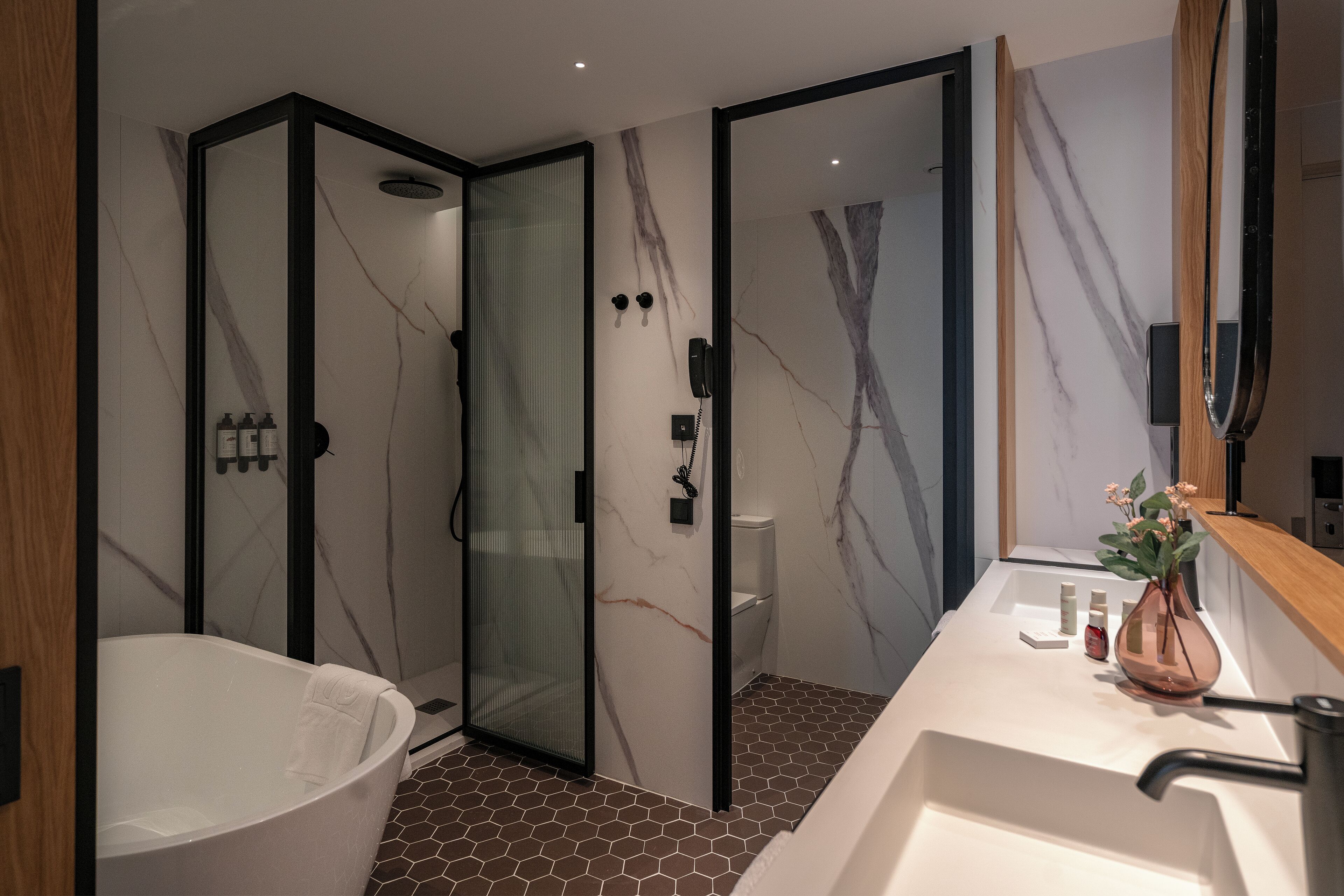 Een moderne badkamer met een douchecabine van glas, een vrijstaand bad en een wastafel met spiegel en een vaas met bloemen.