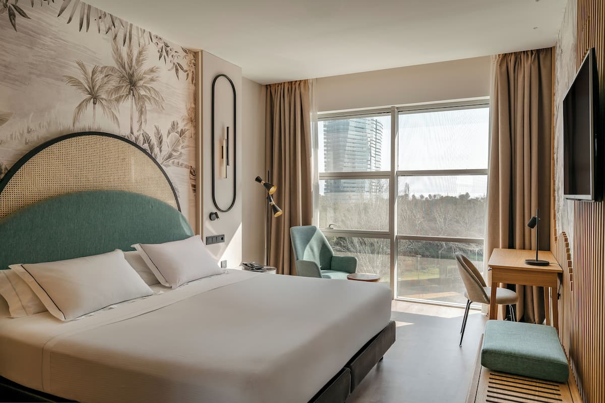 Moderni hotellihuone, jossa on suuri sänky, työpöytä, tuoli ja suuri ikkuna, josta avautuu näkymä kaupunkiin.