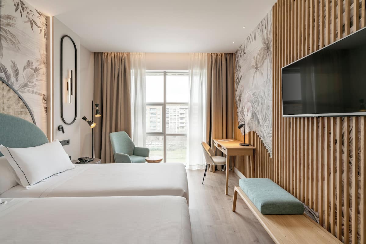 Hotellihuone, jossa on kaksi sänkyä, työpöytä, tuoli, televisio ja parveke, jolta on näkymä rakennuksiin.