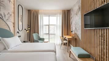 Une chambre d’hôtel avec deux lits, un bureau, une chaise, une télévision et un balcon donnant sur des immeubles.