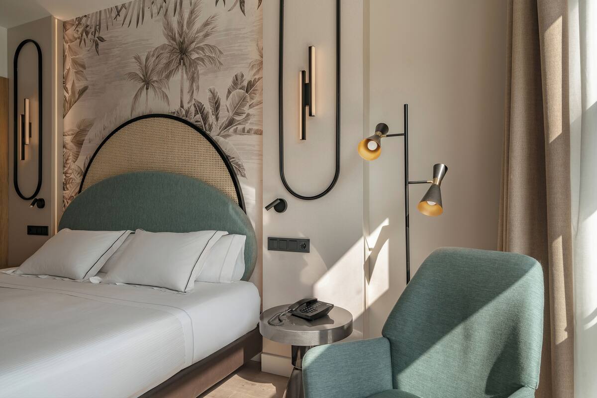 Hotellihuone, jossa on sänky, pyöreä sivupöytä, jossa on puhelin, tuoli, seinään kiinnitetty lamppu ja trooppiseen teemaan sopiva tapetti.