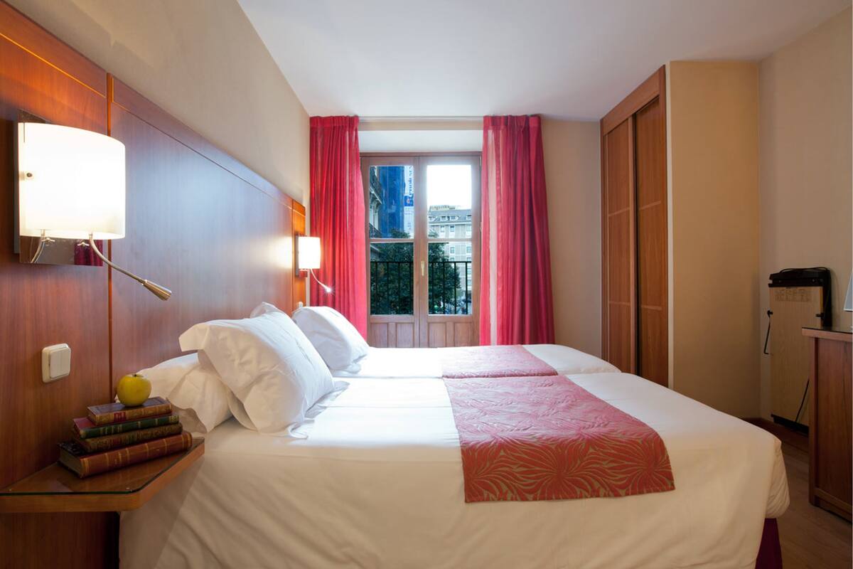 A hotel room with a bed, a nightstand with books, a lamp, and a view of the outside through a window with curtains.