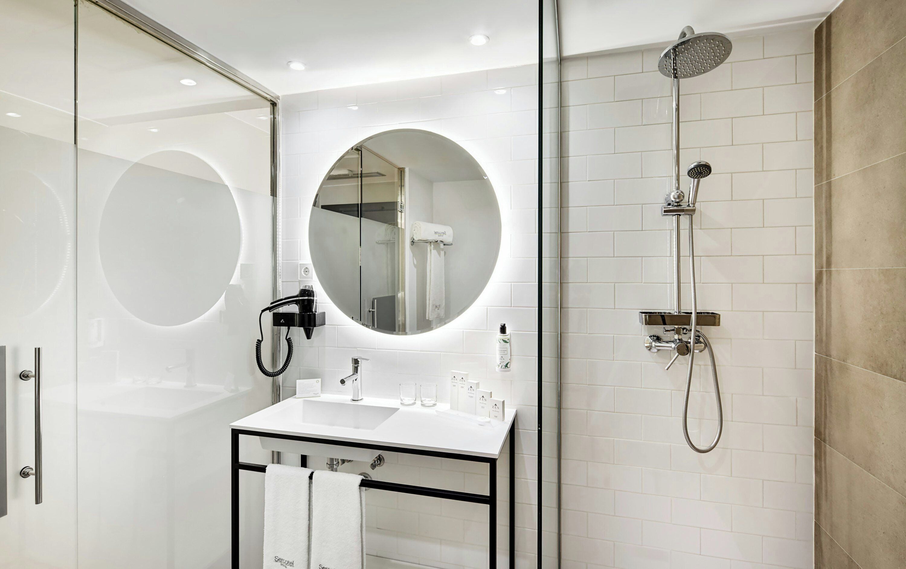 Une salle de bain moderne avec une douche à parois de verre, un miroir rond et un lavabo blanc doté d’un cadre noir.