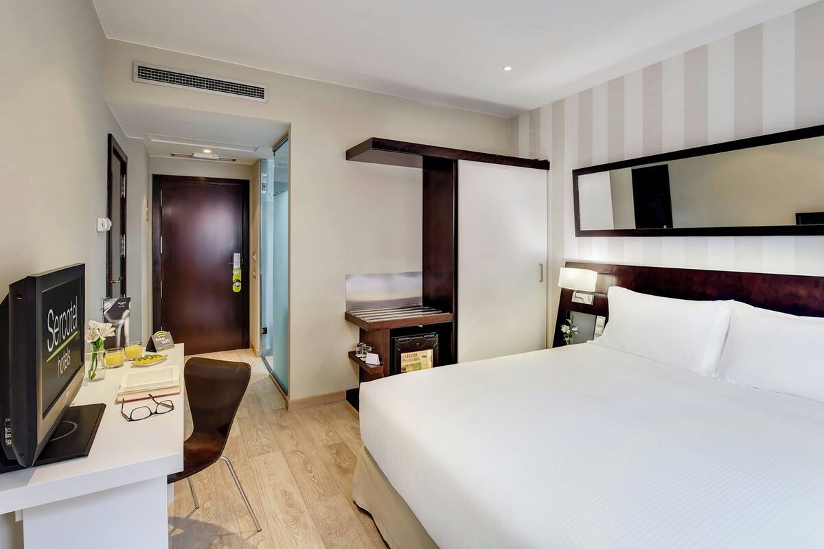 A hotel room with a bed, a desk with a computer, a TV, a minibar, and a mirror.