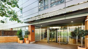 Un bâtiment moderne avec une entrée en verre, des plantes en pot et une enseigne indiquant « Sercotel Madrid Aeropuerto ».