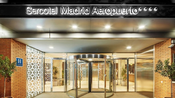L’entrée d’un hôtel, avec une porte tournante, une enseigne au-dessus indiquant « Sercotel Madrid Aeropuerto », et des plantes en pot de chaque côté.