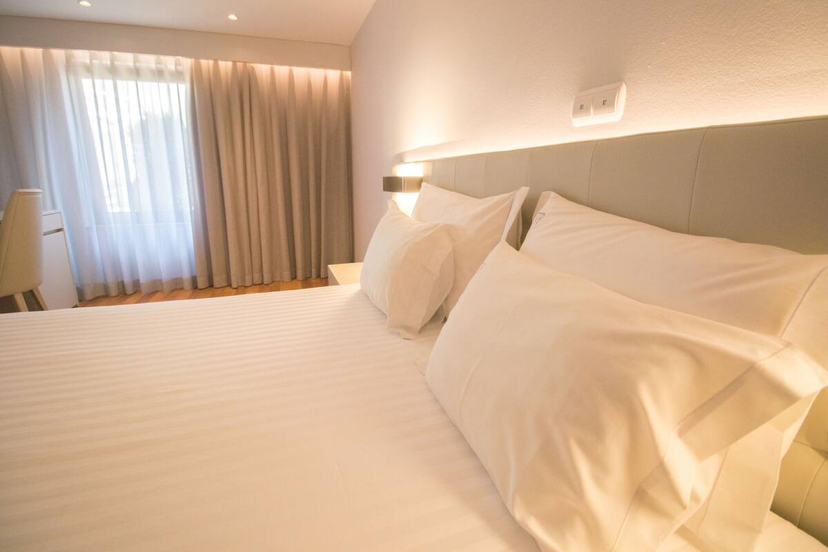 A hotel room with a large bed, a bedside table, a lamp, and a window with curtains.