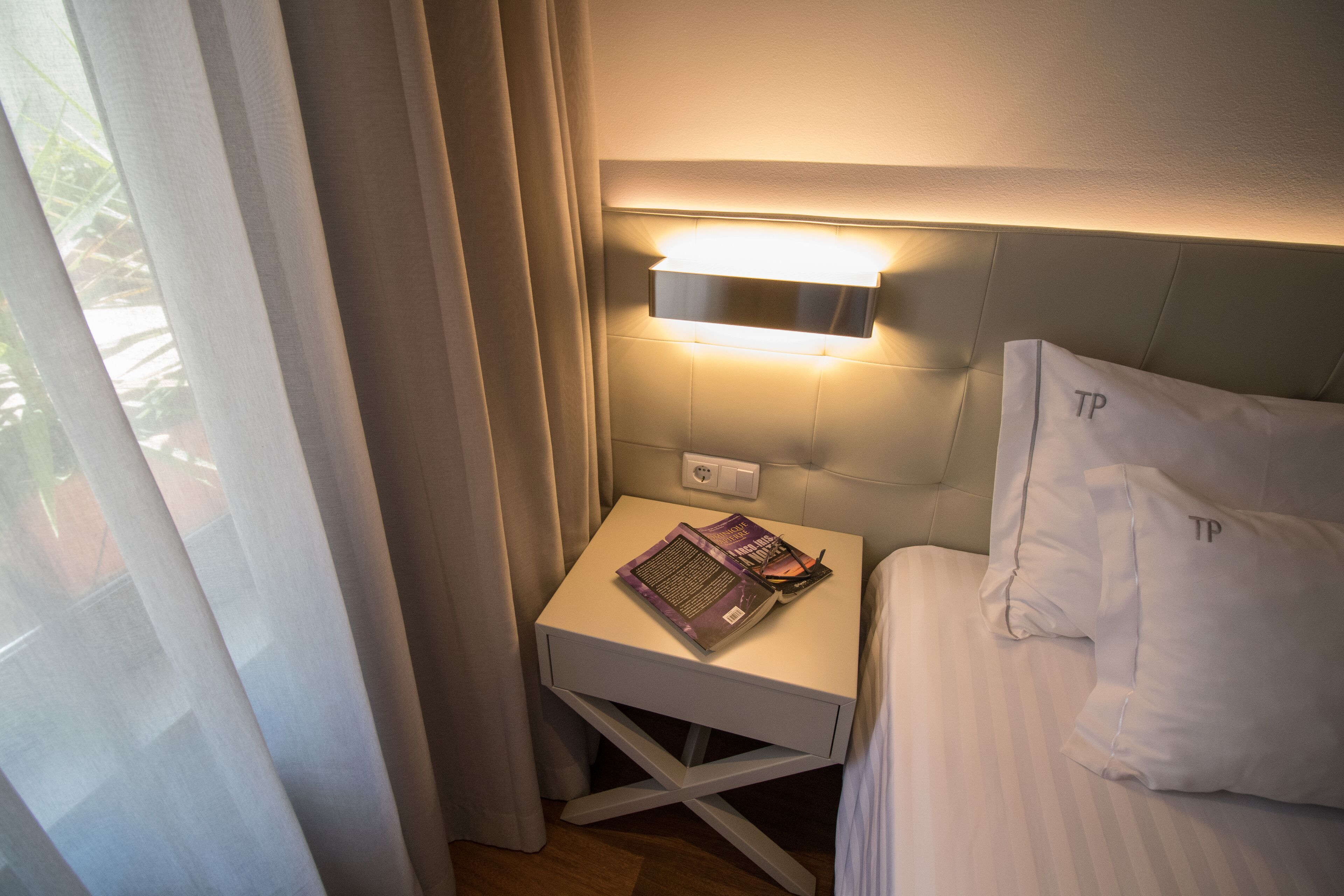 A neatly made bed with white bedding and pillows marked with 'TP'. A small bedside table with a lamp and a book is visible.