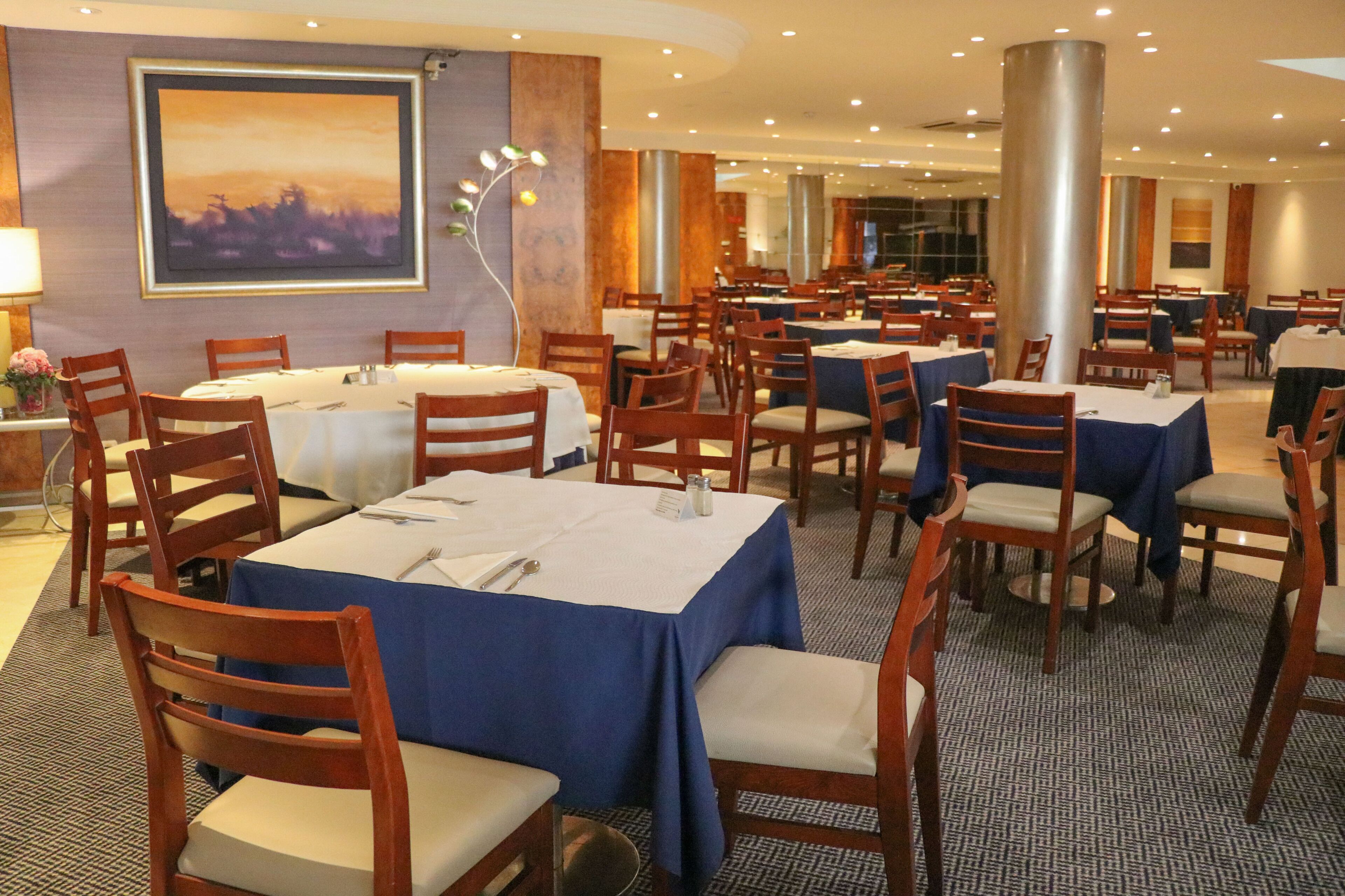 A hotel dining area with multiple round tables, wooden chairs, and a large painting on the wall.