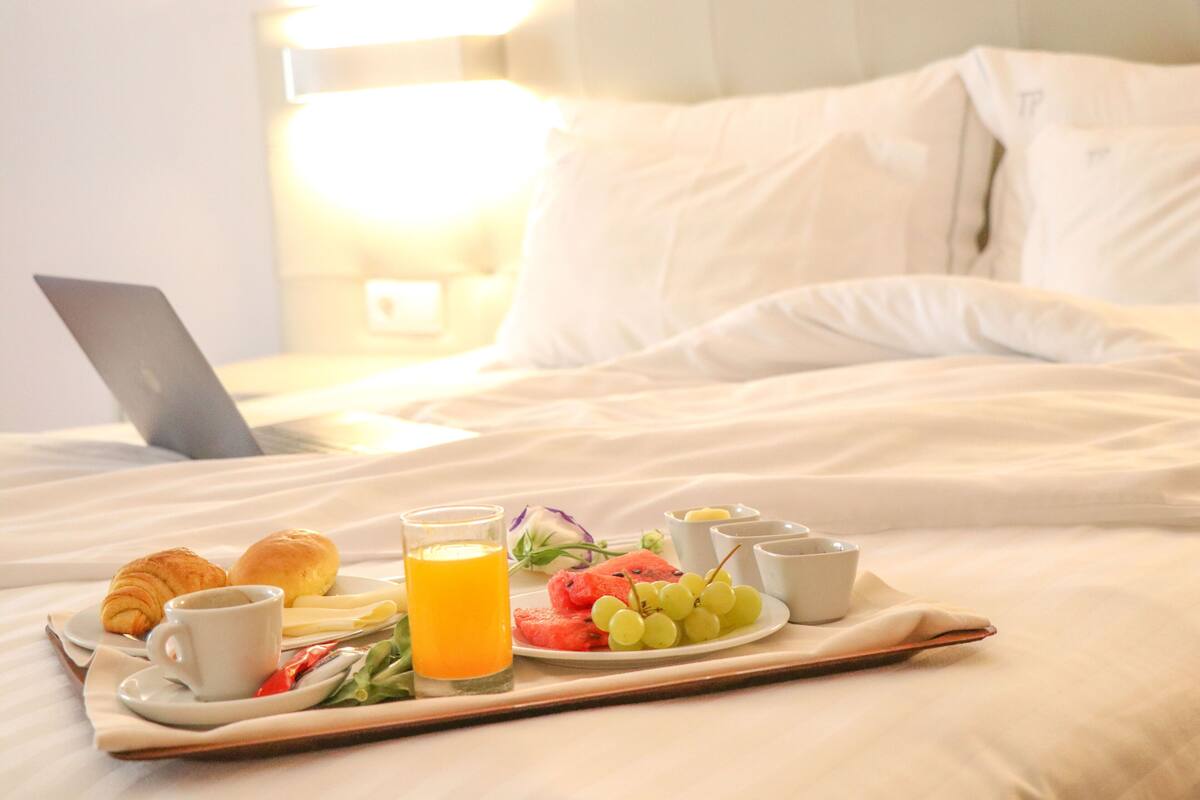 A breakfast tray with croissants, a glass of orange juice, and a plate of fruits on a bed with a laptop and pillows.