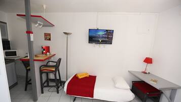 Habitación de hotel pequeña con cama, escritorio y silla, televisión y una mesita de noche con lámpara.