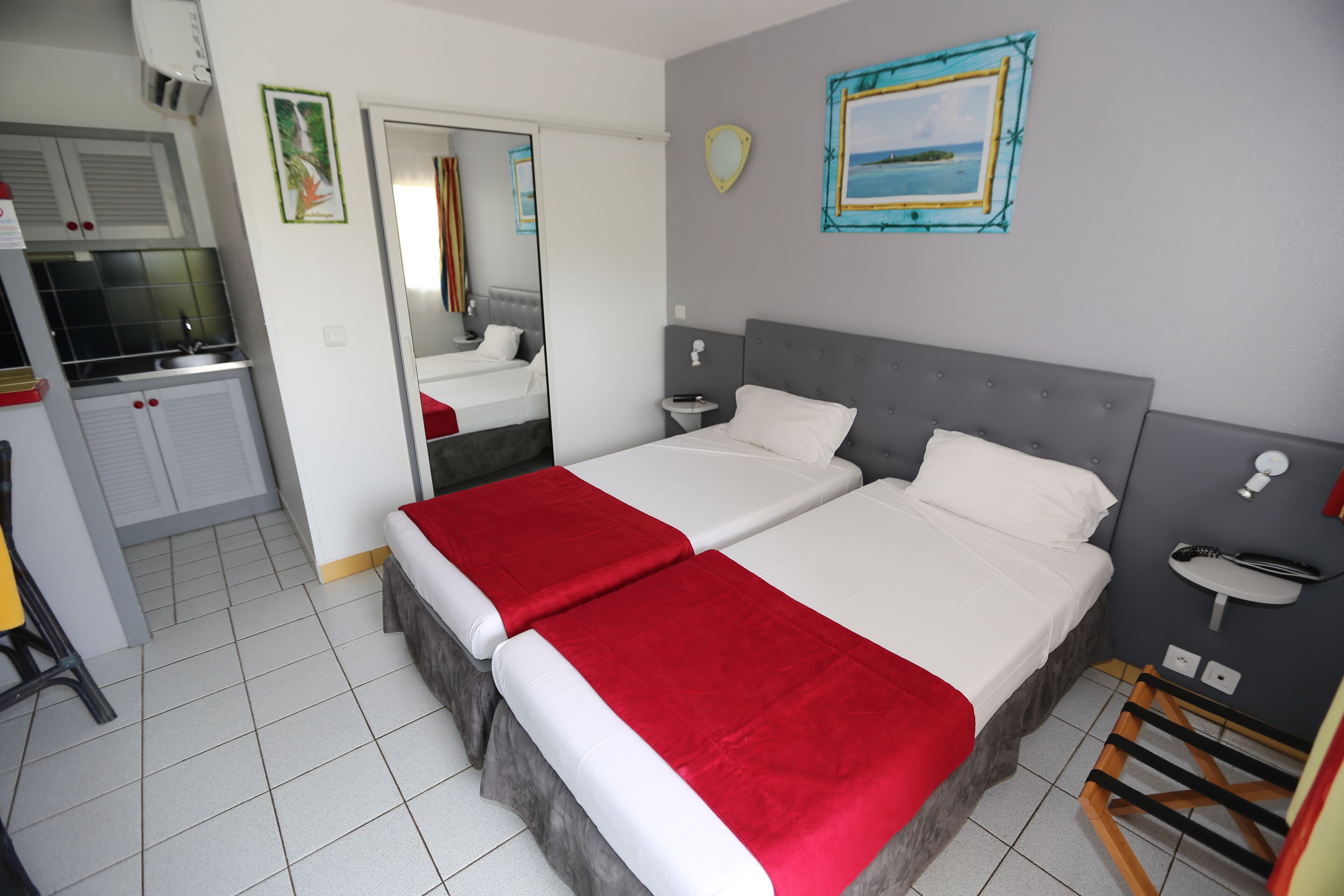 A bedroom with two beds, a small bedside table, a mirror, a wall-mounted picture, and a kitchenette area.
