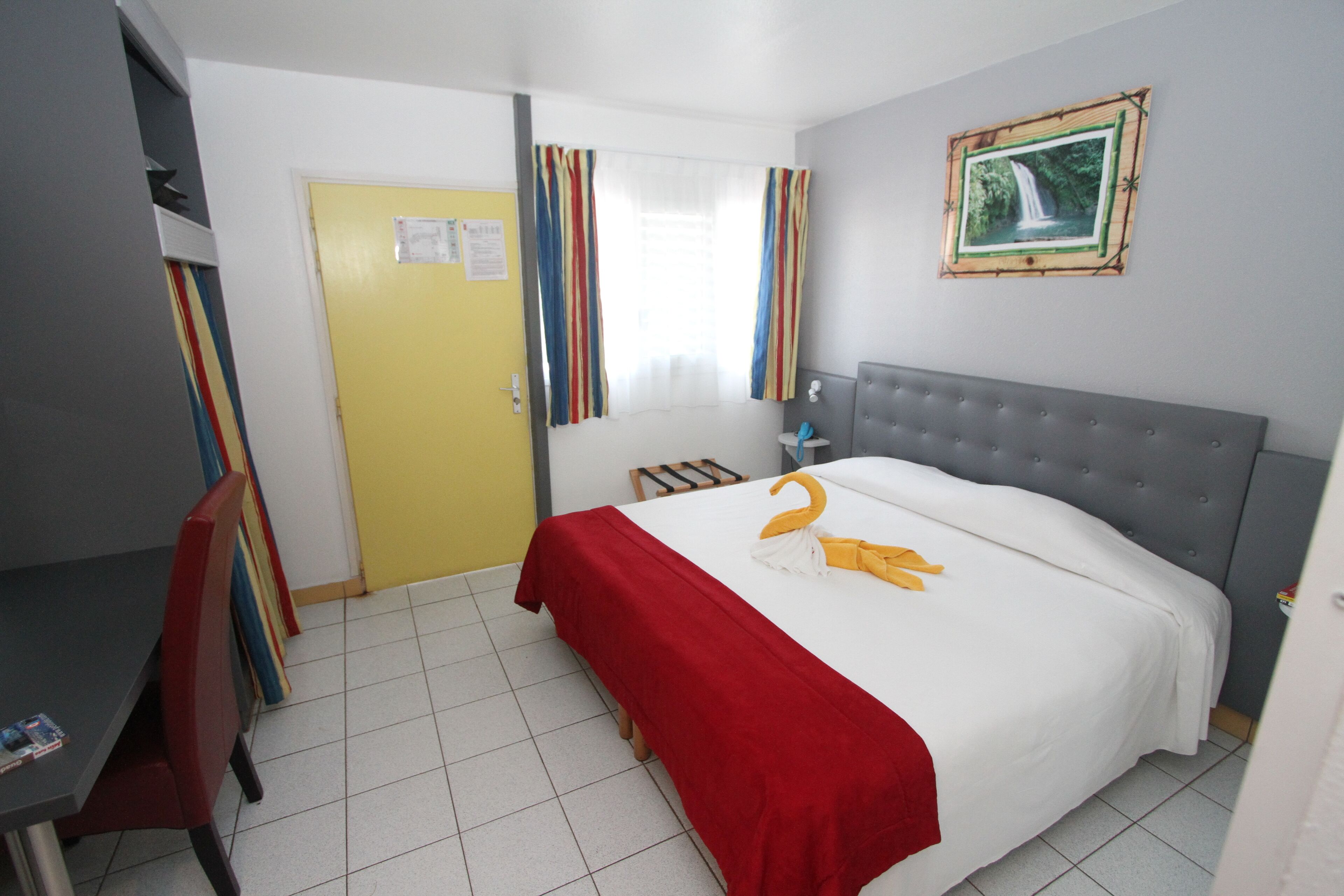 Habitación de hotel con cama, escritorio y silla. Cuenta con una ventana con cortinas, un cuadro en la pared y una puerta.