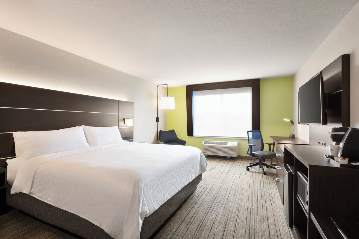A hotel room with a large bed, a desk with a computer, a TV, and a window with blinds.
