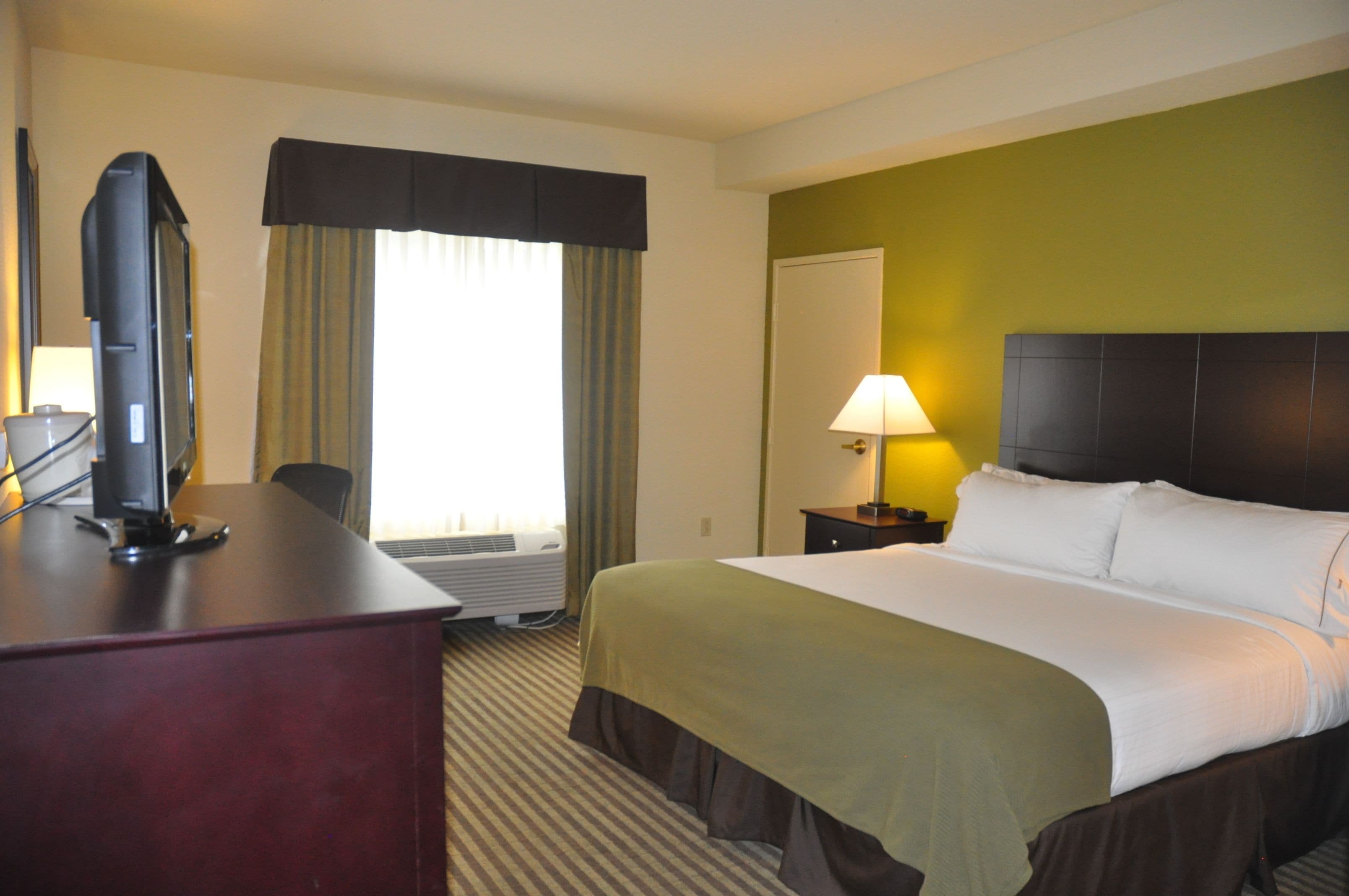 A hotel room with a bed, a desk with a television, a lamp, and a window with curtains.