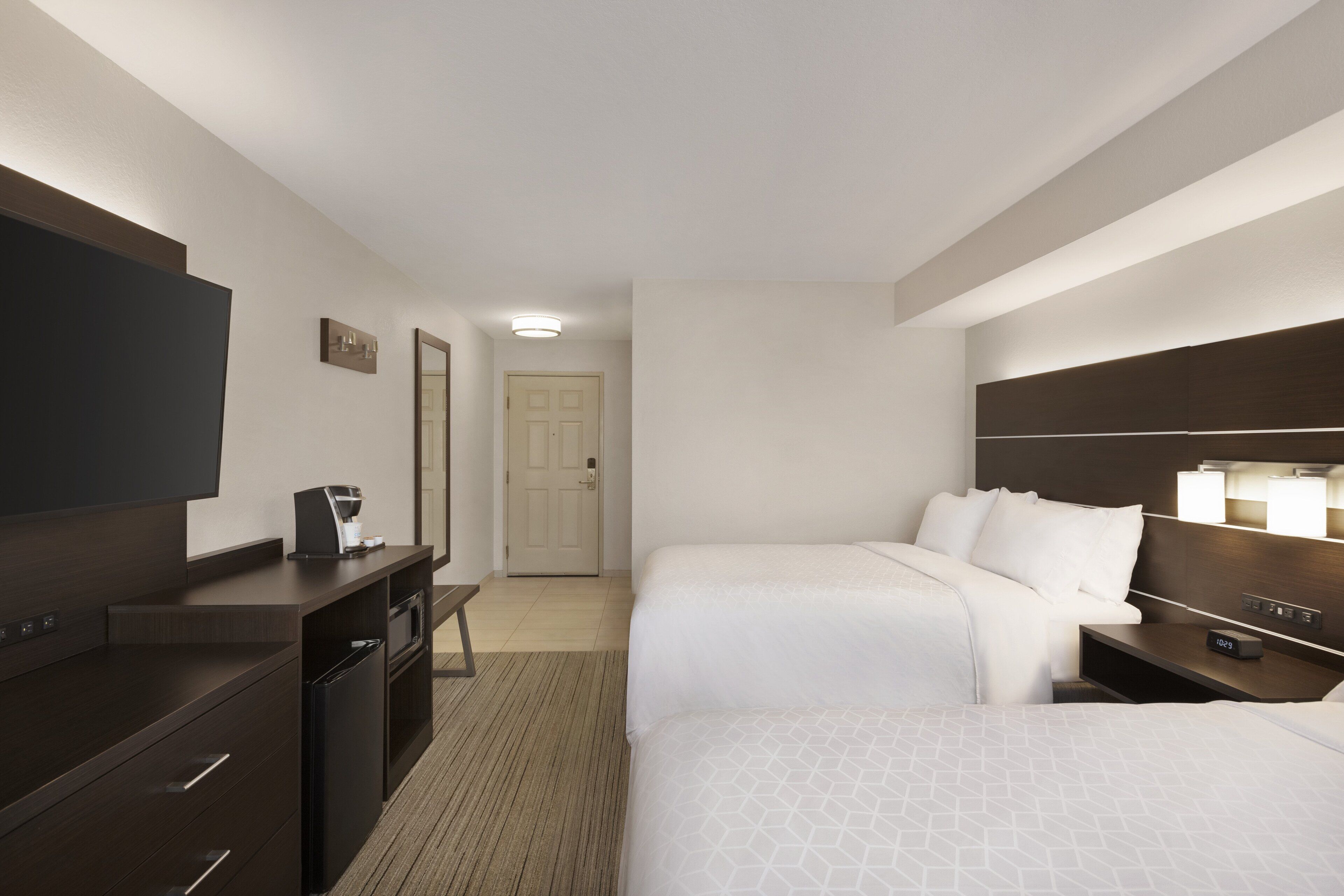 A hotel room with two beds, a desk with a coffee maker, a TV, and a door leading to another room.