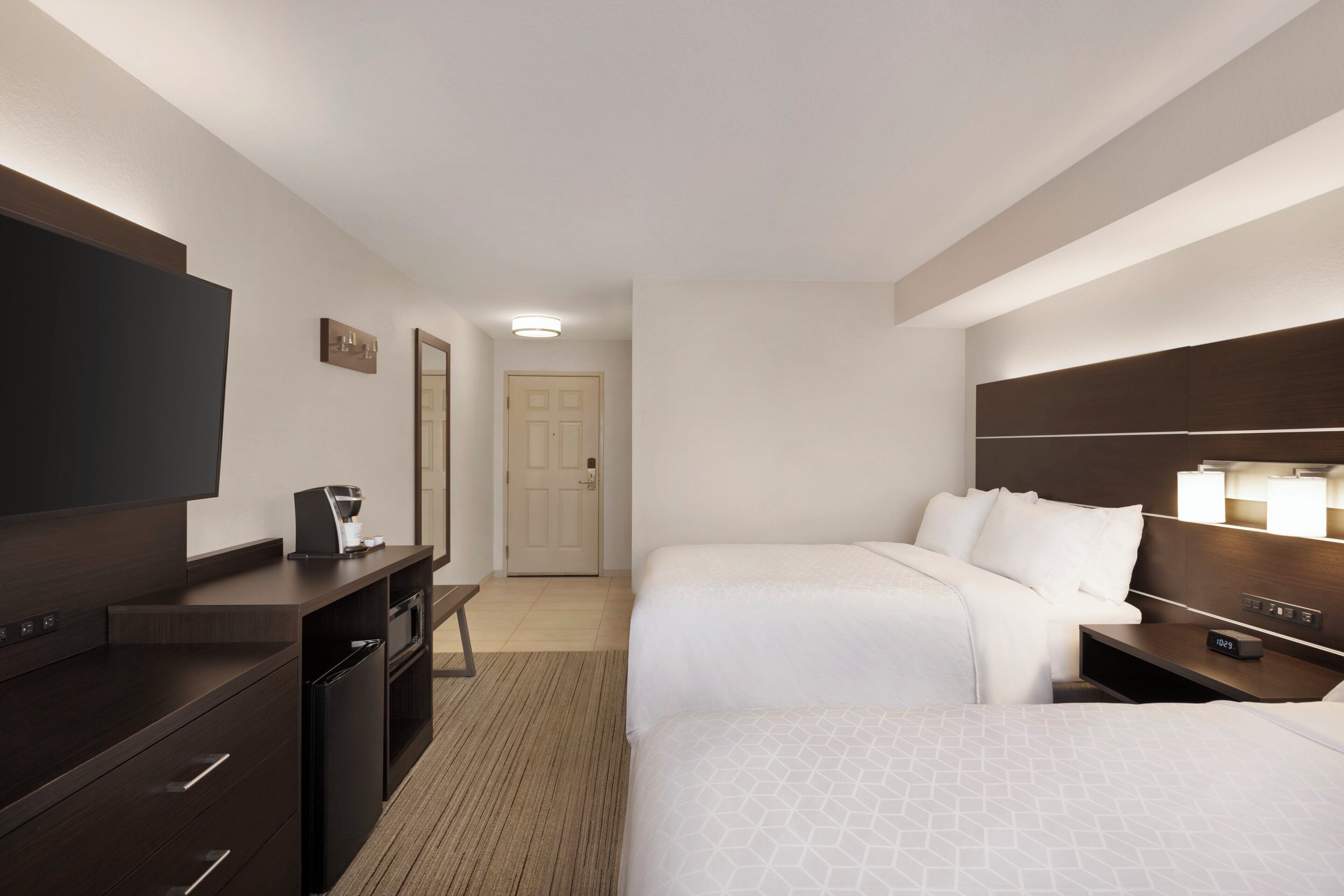 A hotel room with two beds, a desk with a coffee maker, a TV, and a bedside table with a lamp.