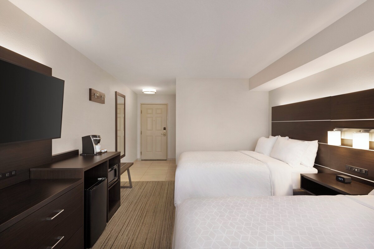 A hotel room with two beds, a desk with a coffee maker, a TV, and a bedside table with a lamp.