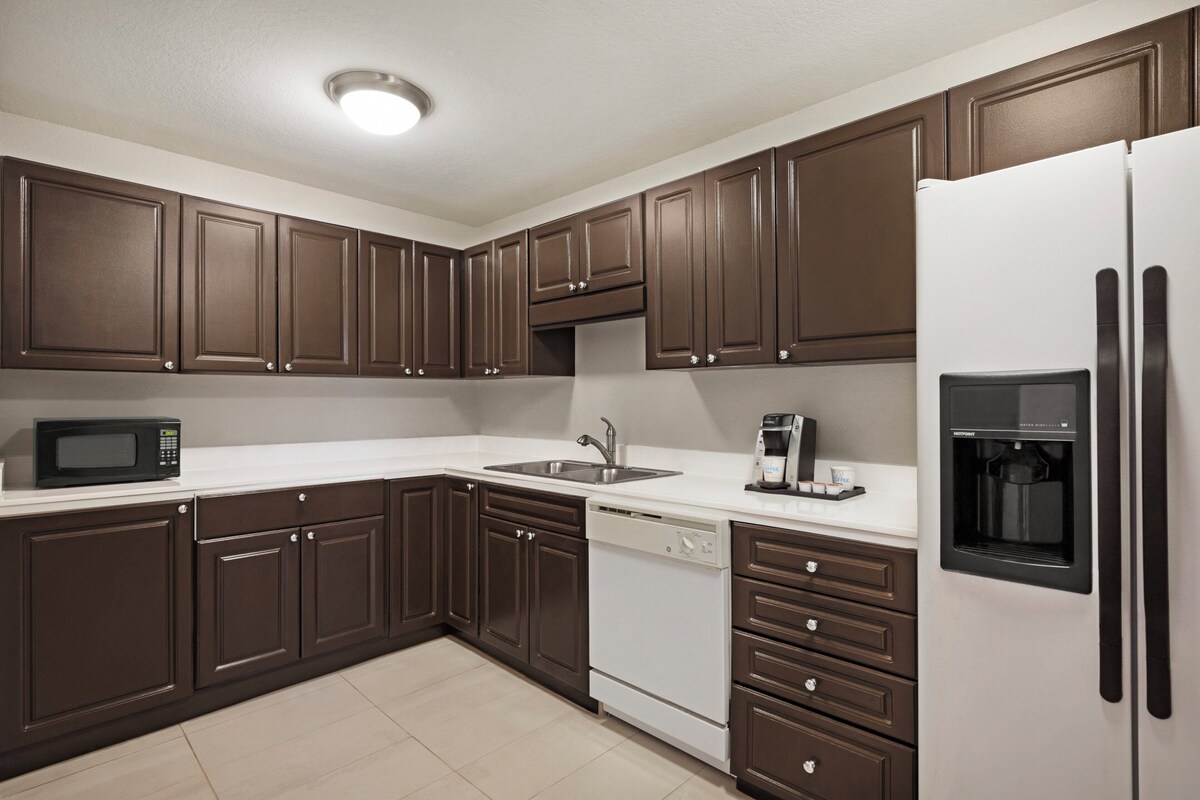 A modern kitchen with dark brown cabinets, a white countertop, a stainless steel sink, a black coffee maker, and a water dispenser.