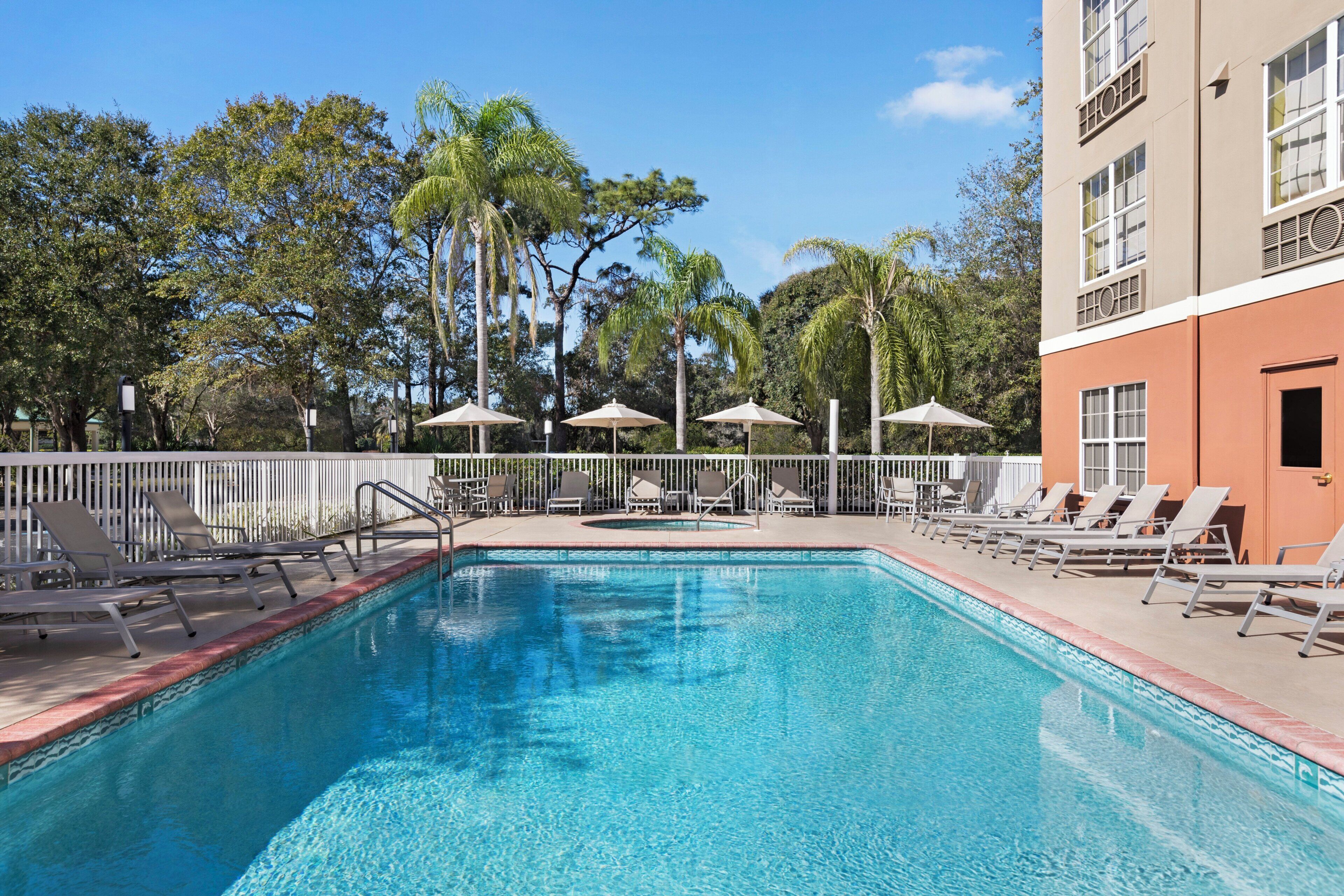 A swimming pool with lounge chairs and umbrellas, surrounded by trees and a building with a 'HOTEL' sign.