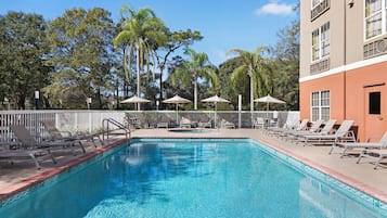 A swimming pool with lounge chairs and umbrellas, surrounded by trees and a building with a 'HOTEL' sign.