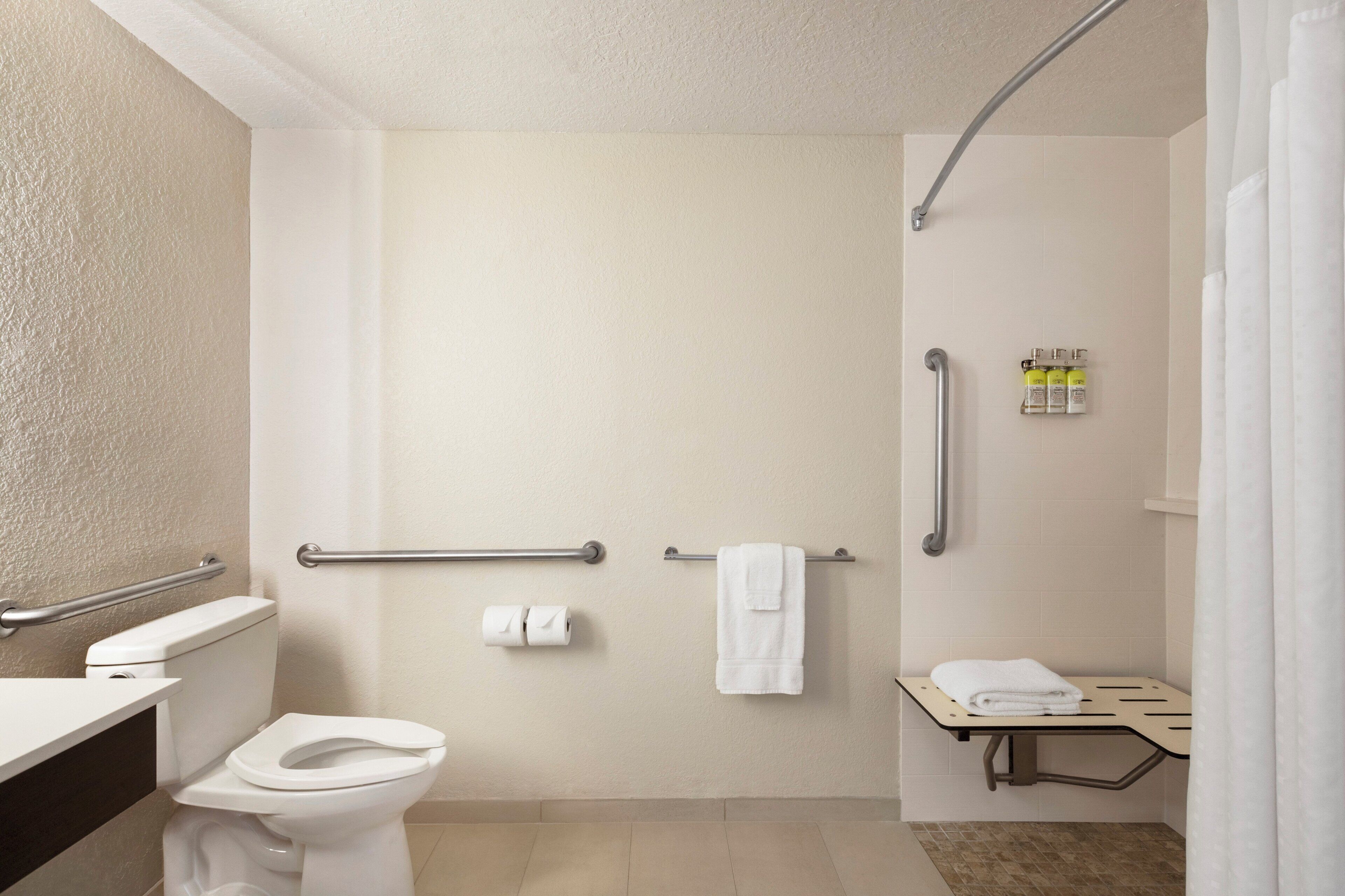 Baño con inodoro, lavamanos, ducha y barra de apoyo.