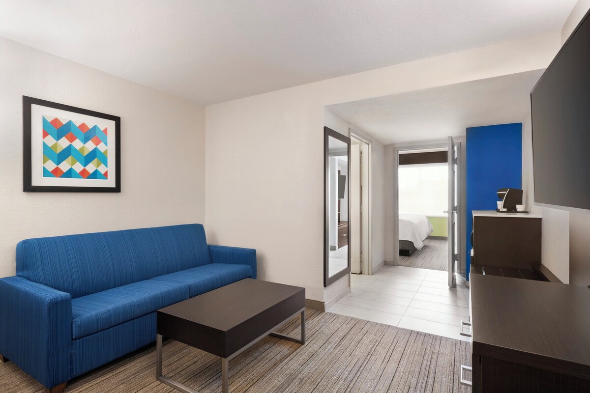 A hotel room with a blue sofa, a small table, a framed picture on the wall, and a view of the adjacent room.