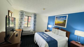 Habitación de hotel con cama, escritorio, silla, ventana con cortinas, un cuadro en la pared y mesita de noche con lámpara.