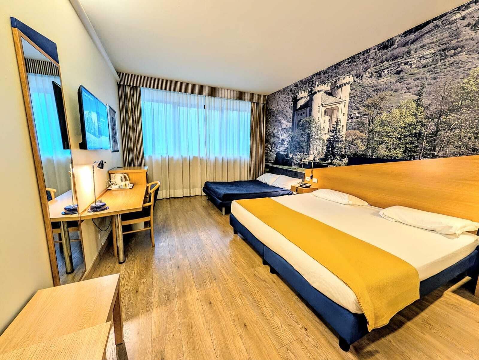 A hotel room with a bed, a desk, a chair, a window with curtains, and a mural on the wall.