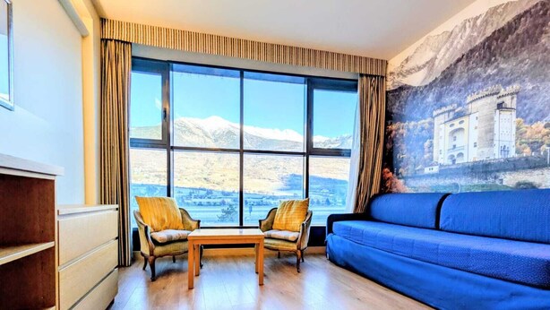 Una habitación con un ventanal grande, un sofá azul, dos sillones, una mesa de madera y un armario.