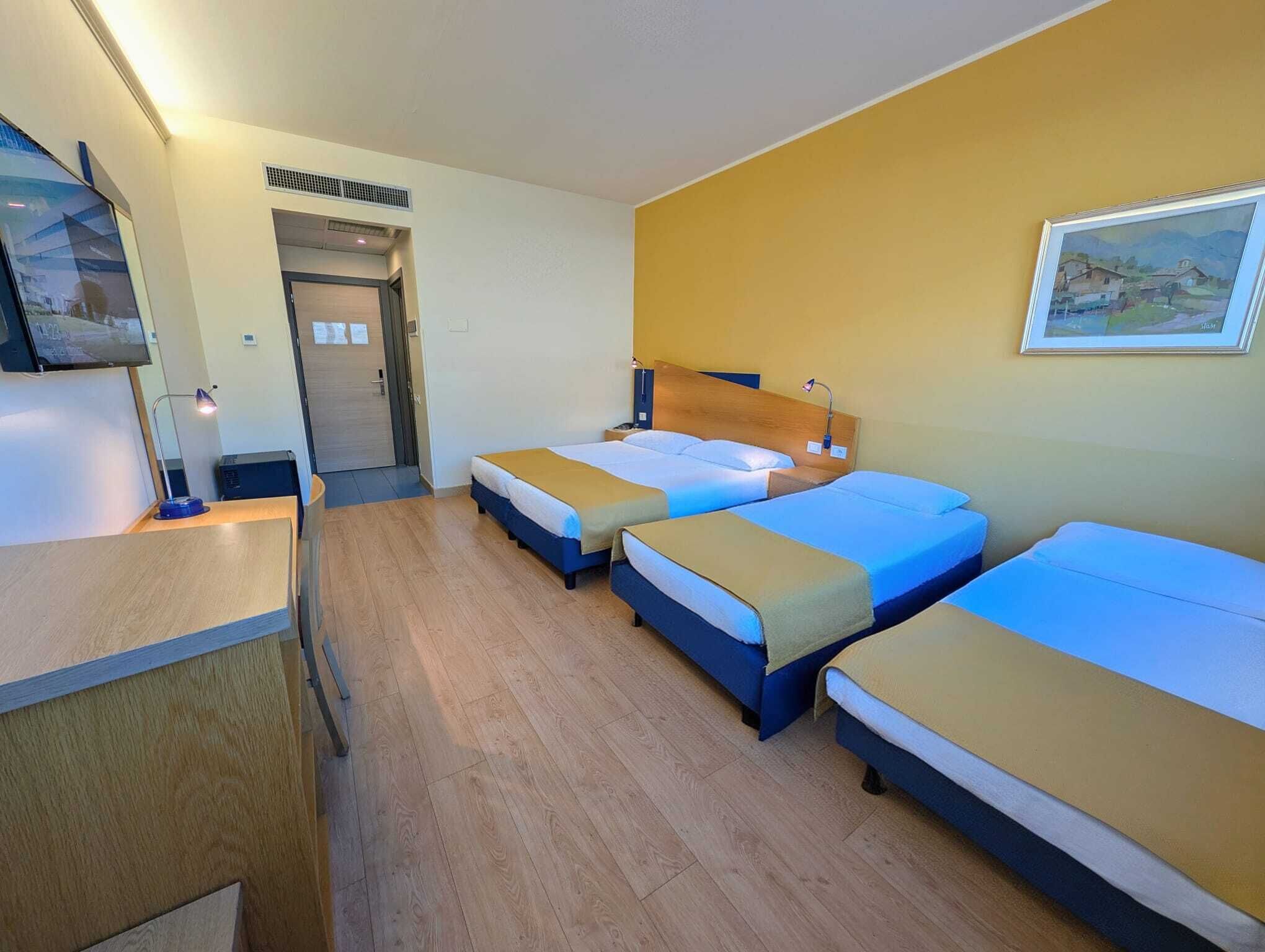 A hotel room with three beds, a desk, a TV, and a painting on the wall.