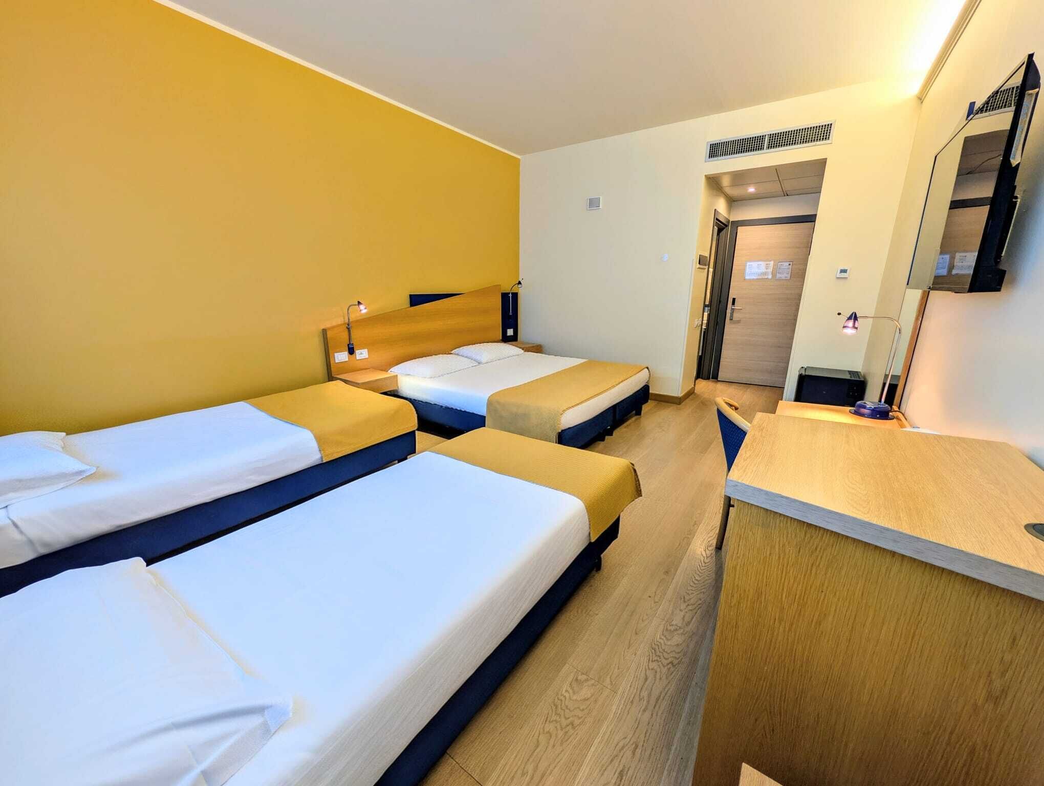 Et hotelværelse med tre enkeltsenge, et skrivebord og et spejl.