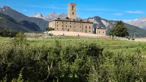 Un edificio de piedra grande con una torre central, rodeado de exuberante vegetación y montañas al fondo.