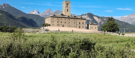 Un edificio de piedra grande con una torre central, rodeado de exuberante vegetación y montañas al fondo.