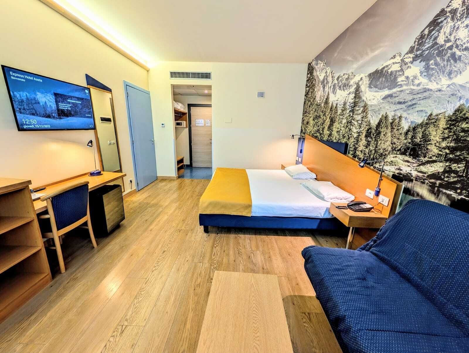A hotel room with a bed, a desk, a chair, a TV, and a mountain view mural.