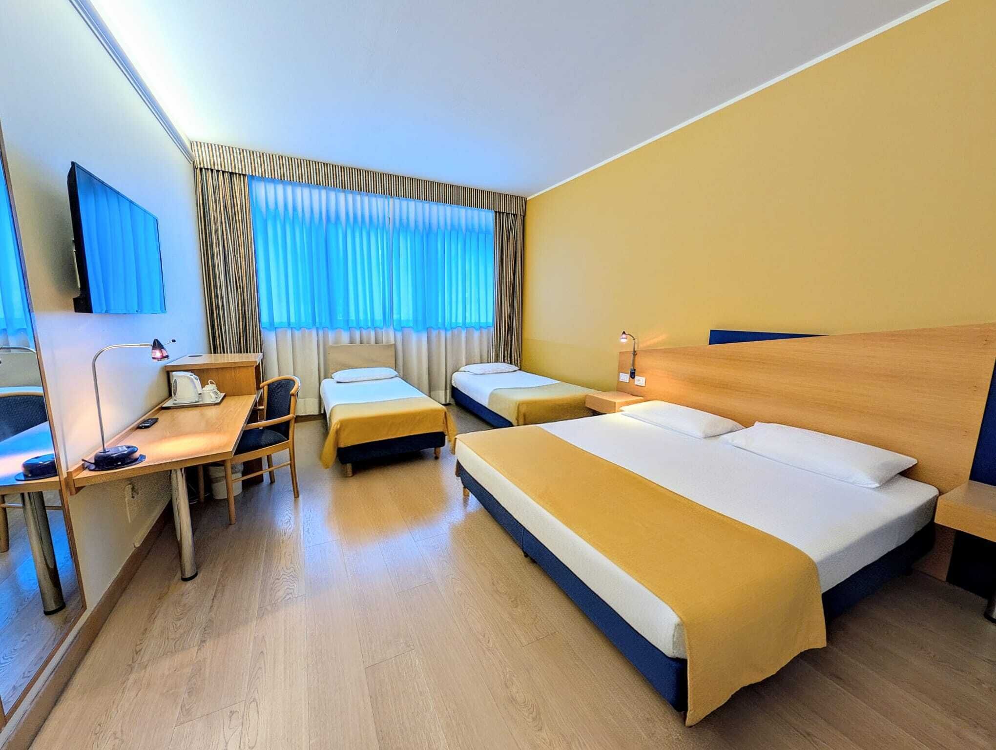 A hotel room with two single beds, a desk with a chair, a TV, and a window with curtains.