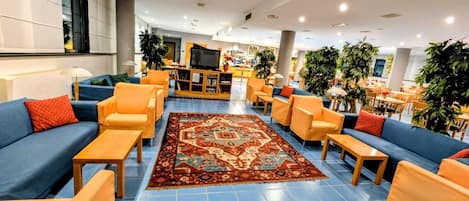 A spacious lobby with blue-tiled flooring, orange and blue sofas, and a central area with a television and bar.