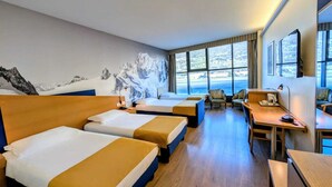 Habitación de hotel con dos camas, un escritorio, una silla y un amplio ventanal con vista a la montaña.