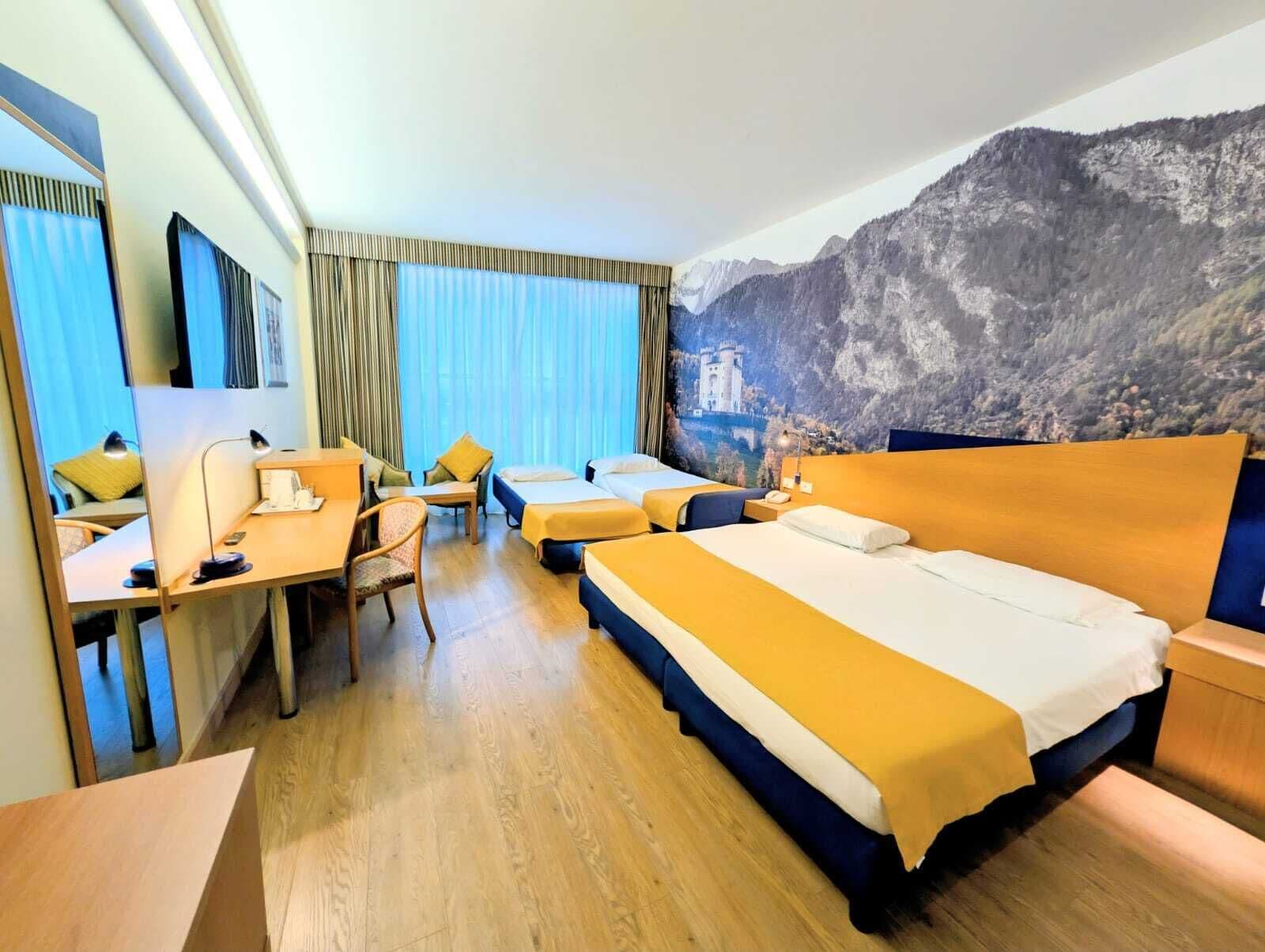 Camera d'albergo con un letto grande, una scrivania e una sedia. Si vede una finestra con tende e un murale raffigurante una montagna sulla parete.