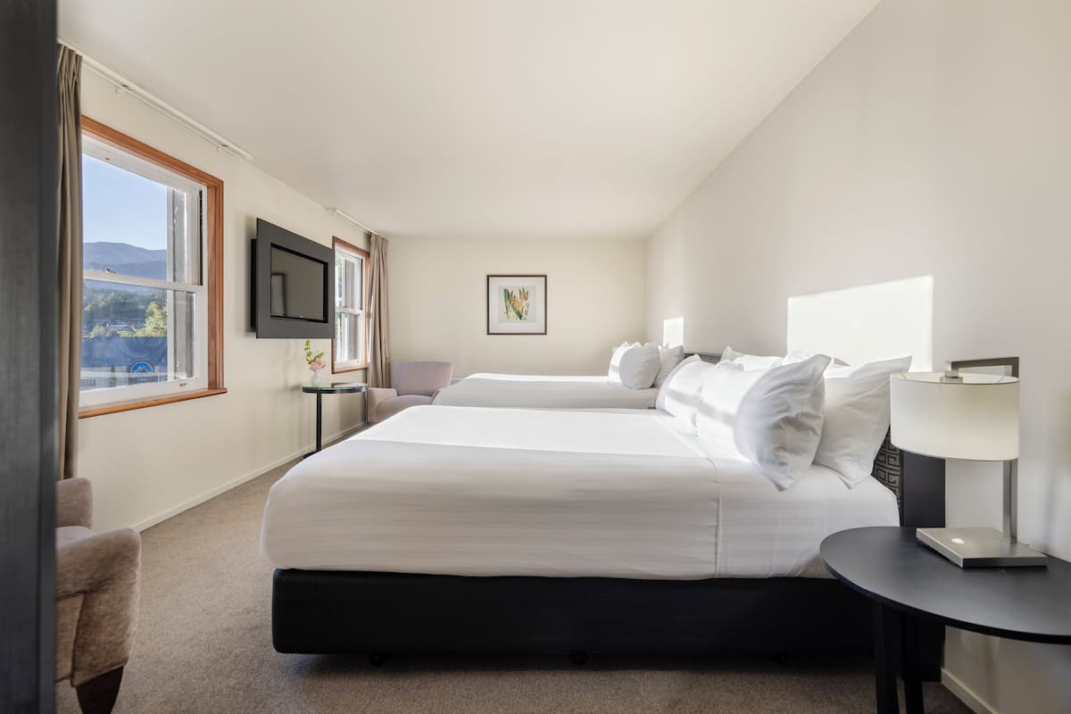 A hotel room with two beds, a TV mounted on the wall, a small table with a lamp, and a window with a view of the mountains.
