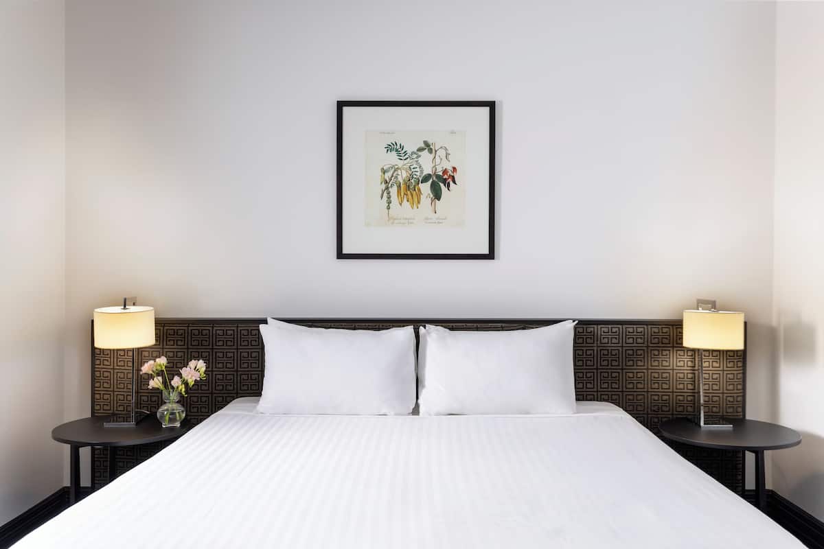 A neatly made bed with white linens, two bedside tables with lamps, and a framed botanical illustration on the wall.
