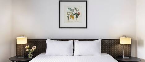Une chambre d’hôtel avec un lit soigneusement fait, deux tables de chevet avec des lampes et une illustration botanique encadrée accrochée au mur.