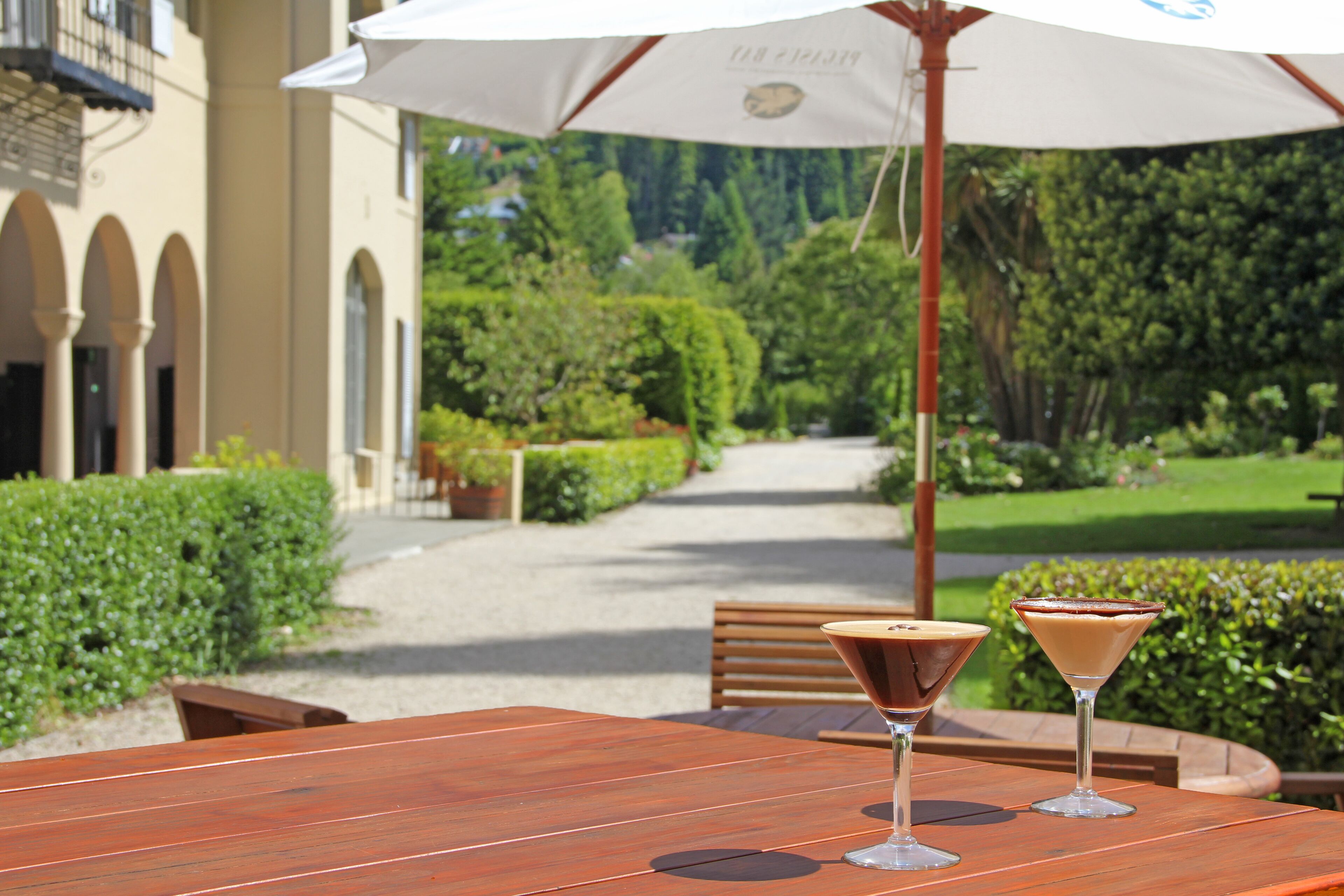 A wooden table with two martini glasses, an umbrella, and a bench in a garden setting.