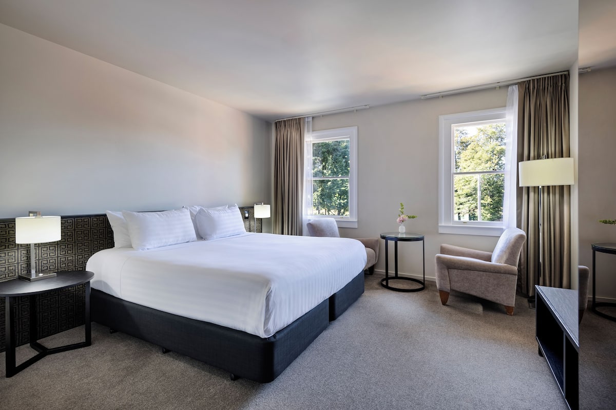 A hotel room with a large bed, two bedside tables, a chair, and two windows with curtains.