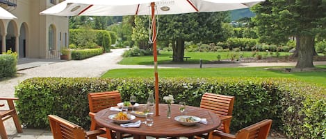 Pyöreä puinen pöytä, jossa on kuusi tuolia, valkoinen aurinkovarjo ja valkoisten kukkien maljakko.