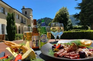 Una mesa con una tabla de embutidos, botellas de cerveza Asahi y Peroni, y una copa de vino blanco, ubicada al aire libre con un edificio y árboles al fondo.