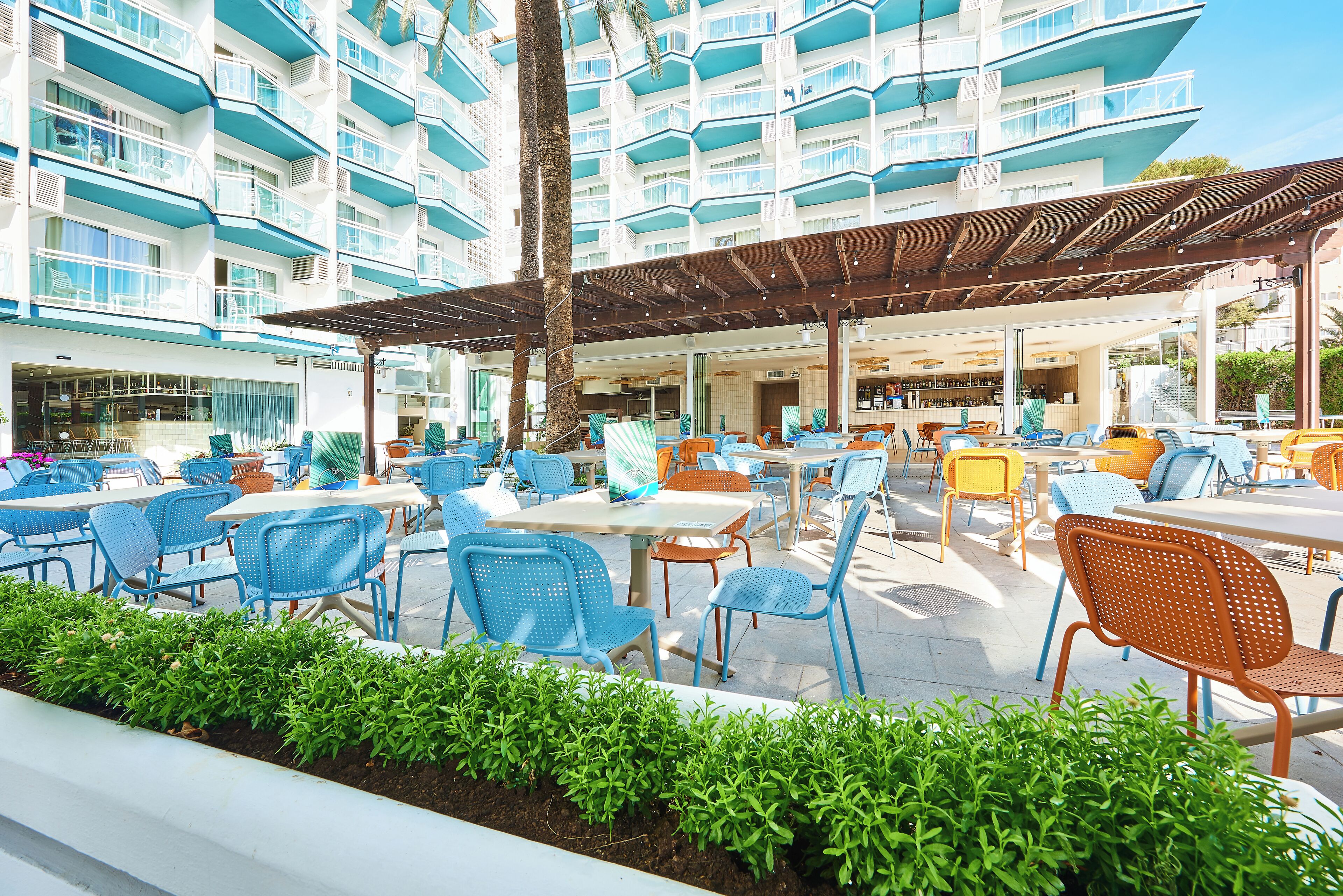 An outdoor seating area with tables and chairs, a covered patio, and a building with multiple balconies in the background.