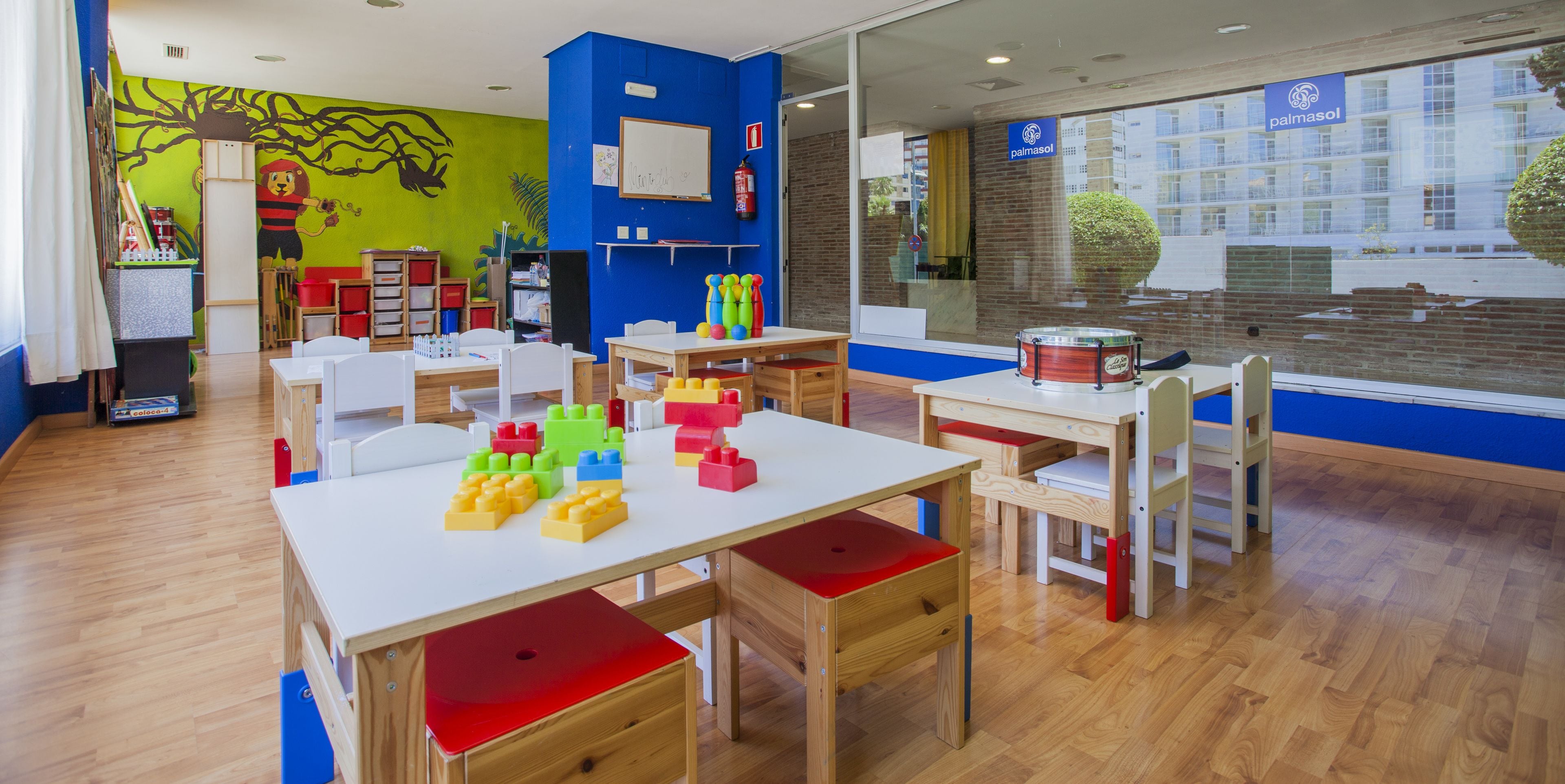 A classroom with wooden tables and chairs, colorful building blocks, and a mural on the wall.